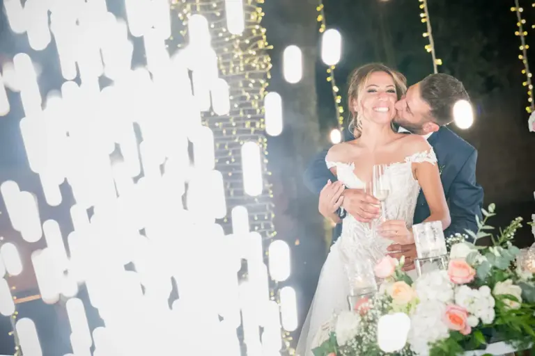 Momento di amore e brindisi nella galleria di Maraca Fotografia: lo sposo felice bacia la sposa tra le fontane fredde della suggestiva location Villa Zaira. Un'immagine che cattura la gioia e la freschezza di questo momento speciale nel giorno del matrimonio.